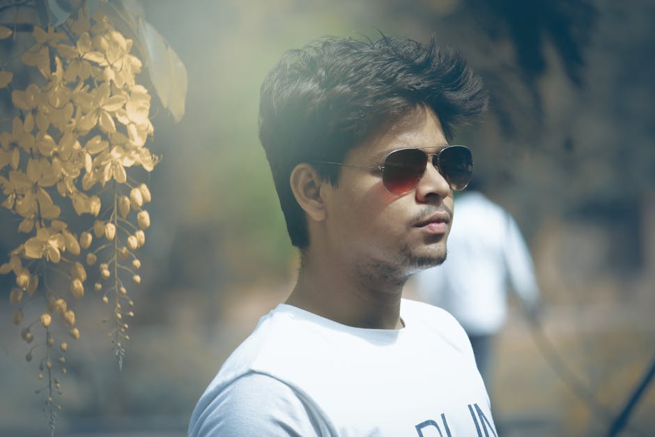 Stylish young man in sunglasses poses outdoors against a backdrop of yellow flowers, embodying a casual fashion vibe.